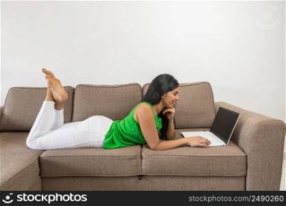Side view of barefoot Hispanic female in casual clothes leaning on hand and browsing netbook with black screen while resting on couch in living room at home. Hispanic woman using laptop on sofa