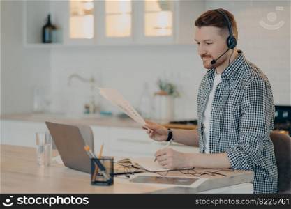 Side view of bearded man making notes in notebook and holding papers with business data while using headset during online call on laptop, sitting at his cozy workplace in kitchen. Remote work at home. Smiling guy during online meeting while working at home