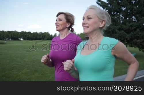 Side view of beautiful adult blonde girlfriends running in park road. Closeup. Angle view. Positve running senior females jogging during outdoor workout in park. Slow motion. Steadicam stabilized shot.