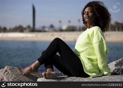 Side view of beautiful curly afro woman sitting on breakwater rocks laughing while looking camera outside