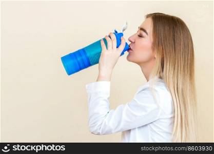 Side view of beautiful young caucasian woman is drinking water from plastic bottle against a beige background. Woman drinking water from bottle