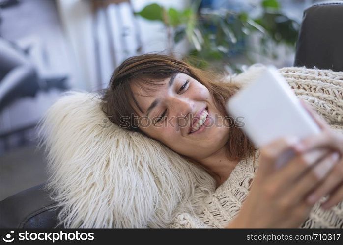 side view of beautiful young smiling woman lying on leather couch relaxing while using a mobile phone at home. Rafa Fernandez