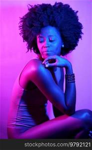 Side view of black woman in bodysuit with Afro hairstyle touching chin and looking away over shoulder while sitting under neon light against purple background. African American model under neon illumination