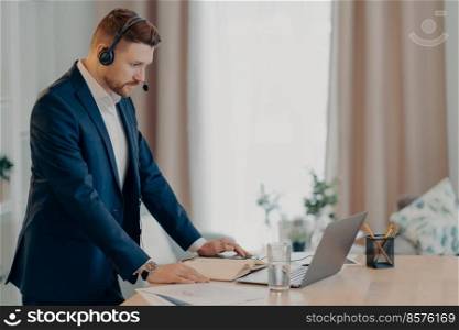 Side view of business consultant wearing suit and headset working on laptop during online call, serious and concentrated businessman surfing internet while standing indoors. Remote work concept. Young businessman standing in room with too much work online
