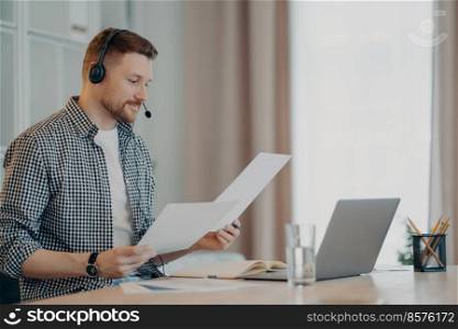 Side view of busy young man working with laptop and using headset while having online meeting. Male freelancer analyzing project documents or sales report at his workplace. Remote job at home. Smiling bearded guy looking at documents while working from home