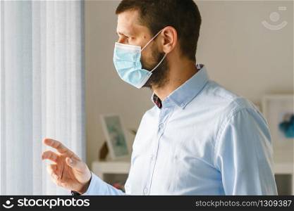 Side view of caucasian man standing by the window wearing medical protective mask to protect from virus bacteria pandemic disease while working from home quarantine or office looking out wearing shirt