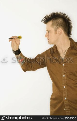 Side view of Caucasian man with mohawk holding and aiming dart against white background.