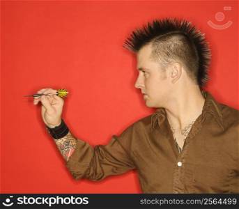 Side view of Caucasian man with mohawk holding dart against red background.