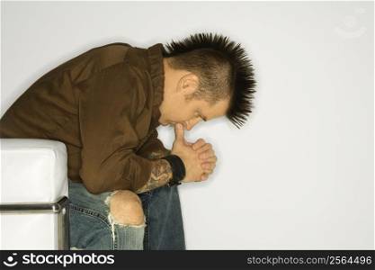 Side view of Caucasian man with mohawk sitting with hands to face against white background.