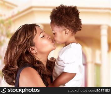 Side view of cheerful mother holding on hands cute little curly son and kiss him, spending time outdoors near great beautiful house, happy African family