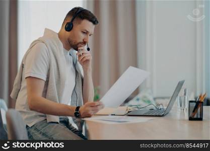 Side view of concentrated bearded man listening attentively to online lesson while studying remotely at home, holding document in hand and analyzing information. Online study concept. Young guy in headphones holding paper and looking at laptop screen at home
