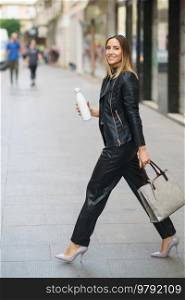 Side view of confident stylish woman in leather outfit and heels carrying modern handbag with eco friendly bottle and walking in city. Modern trendy female with reusable bottle in downtown