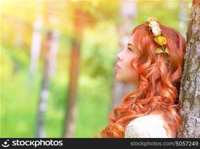 Side view of cute romantic woman looking on sun rays, wearing gentle wreath on curly red hair, fashionable photoshoot in the park