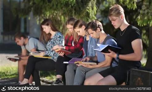 Side view of diverse college students sitting on the bench in park, reading books and textbooks while studying together on campus. Group of university friends learning together outdoors while preparing for final high school exams.