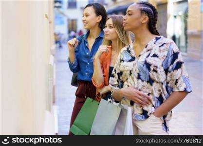 Side view of diverse female friends with shopping bags looking at showcase while choosing goods together on street of city. Multiracial women standing near showcase