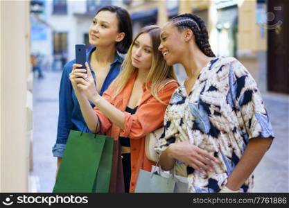 Side view of diverse female friends with shopping bags taking picture of showcase while standing near store on street during shopping. Multiethnic women taking photo of showcase while choosing goods