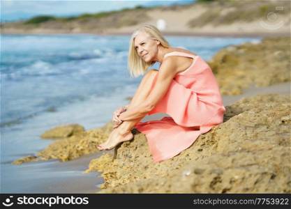 Side view of dreamy graceful elderly female in dress sitting on stone and admiring sea on sunny day in summer. Slim senior woman on rocky seashore in summer