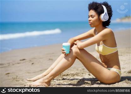 Side view of dreamy young African American female tourist with Afro hair, in bikini drinking takeaway coffee and looking away while listening to music in headphones on sandy seashore. Black woman in headphones drinking takeaway beverage while relaxing on beach