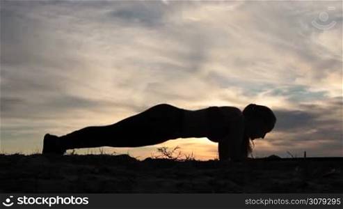 Side view of fitness young female in sport top and leggings doing plank hip twists exercise on beach at sunset. Silhouette of beautiful fiit woman doing plank position looking forward over colorful sky in glow of sunset background.