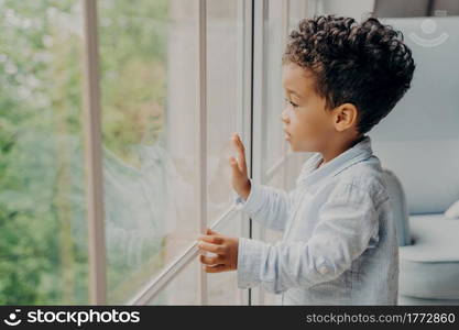 Side view of focused adorable sweet kid boy looking outside through window with surprised and intrigued expression, interested in whats going out there, little child waiting for parents at home. Sweet african american kid looking through window and waiting for parents