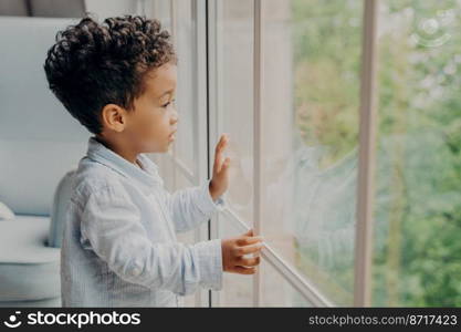 Side view of focused adorable sweet kid boy looking outside through window with surprised and intrigued expression, interested in whats going out there, little child waiting for parents at home. Sweet african american kid looking through window and waiting for parents