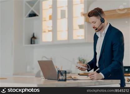 Side view of focused bearded businessman in formal wear working remotely at home, standing indoors and holding sales report, writing notes during an online conference with colleagues. Executive director reading document and writing notes in notepad while working at home