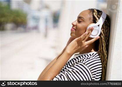 Side view of glad African American female with closed eyes touching headphones and listening to music while leaning on wall on sunny day on blurred background of city street. Optimistic black woman listening to music in city