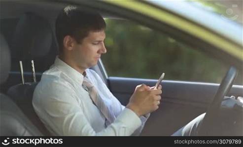 Side view of handsome entrepreneur in formal wear sitting in driver&acute;s seat and surfing the net with smartphone. Succesful businessman browsing the net on mobile phone while driving car during business trip.
