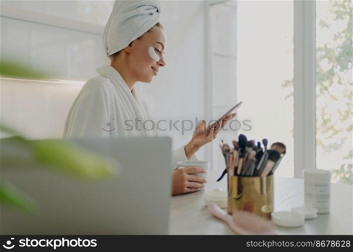 Side view of happy beautiful business woman in bathrobe with hair wrapped in towel and cosmetic patches under eyes using smartphone while getting ready for work in morning at home, standing in kitchen. Beautiful woman in bathrobe with towel on head using smartphone while doing skin care procedures at home