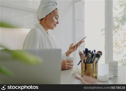 Side view of happy beautiful business woman in bathrobe with hair wrapped in towel and cosmetic patches under eyes using smartphone while getting ready for work in morning at home, standing in kitchen. Beautiful woman in bathrobe with towel on head using smartphone while doing skin care procedures at home