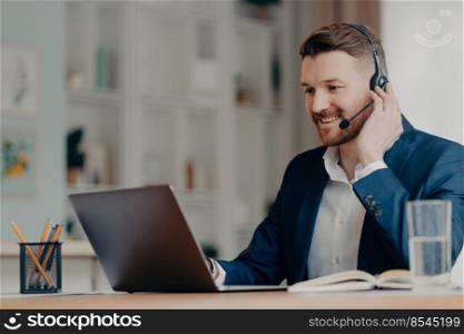 Side view of happy businessman wearing suit using laptop and headset while taking part in online meeting, looking at screen and having web conference with team online. Remote job and freelance concept. Smiling young manager sitting at desk at home and taking part in online meeting