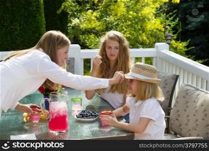 Side view of happy mother putting hat on younger daughter while eating breakfast outdoors, during summer time, on patio with woods in background