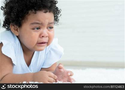 Side view of little sad African American chubby kid girl is crying with tears rolled down her cheeks while lying on fluffy carpet on floor at home. Child emotion care concept. White background