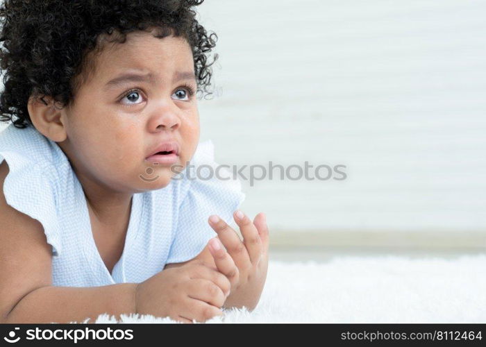 Side view of little sad African American chubby kid girl is crying with tears rolled down her cheeks while lying on fluffy carpet on floor at home. Child emotion care concept. White background