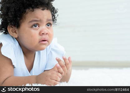 Side view of little sad African American chubby kid girl is crying with tears rolled down her cheeks while lying on fluffy carpet on floor at home. Child emotion care concept. White background