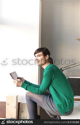 Side view of mature woman sitting on desk holding digital tablet looking away smiling