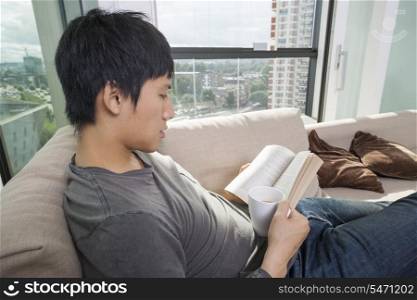 Side view of mid adult man having coffee while reading book on sofa