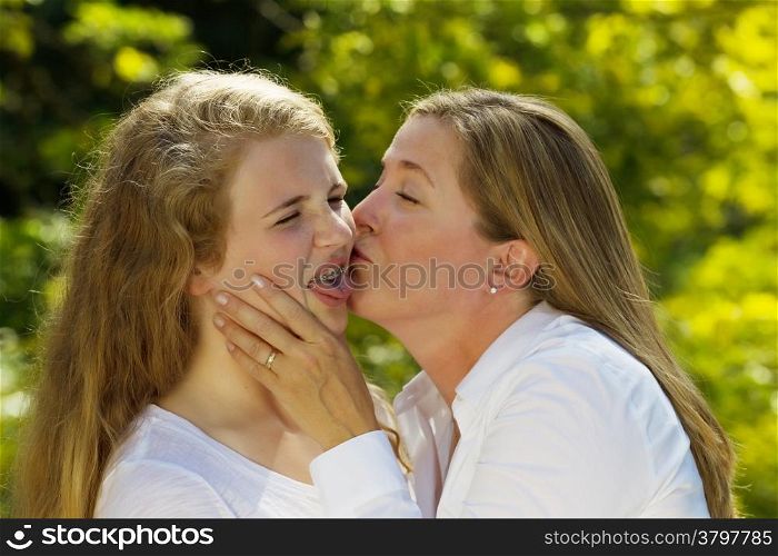 Side view of mother kissing her teenage daughter, expressing herself in a negative way, while holding her face in her hands during outing outdoors on patio with blurred out woods in background