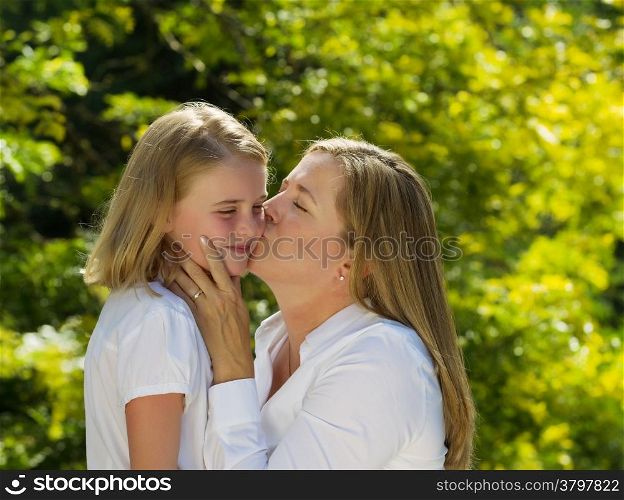 Side view of mother kissing her youngest daughter while holding her face in her hands during outing outdoors on patio with blurred out woods in background