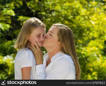 Side view of mother kissing her youngest daughter while holding her face in her hands during outing outdoors on patio with blurred out woods in background
