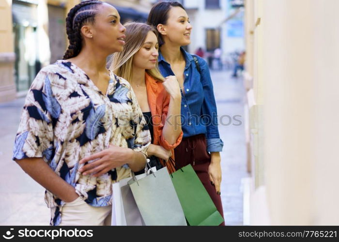 Side view of multiracial female friends with colorful shopping bags standing near boutique while observing showcase on street of city. Multiethnic women standing near store window