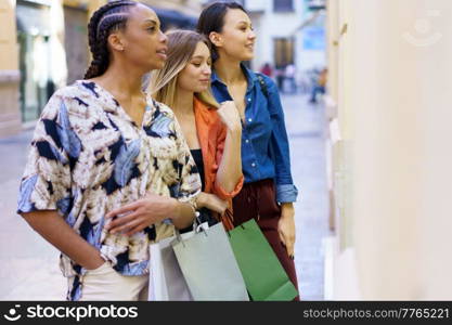 Side view of multiracial female friends with colorful shopping bags standing near boutique while observing showcase on street of city. Multiethnic women standing near store window