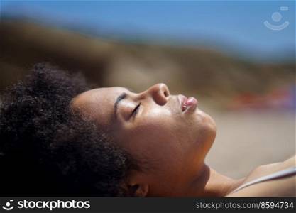 Side view of peaceful black female tourist with Afro dark hair, relaxing on sandy beach with closed eyes, and sunbathing on sunny day during summer holiday. Young African American woman lying on beach with closed eyes