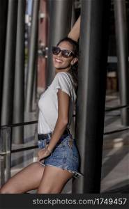 Side view of playful beautiful woman in sunglasses and shorts leaning on pillar while standing at urban terrace on sunny day. Flirty woman in sunglasses leaning on street column