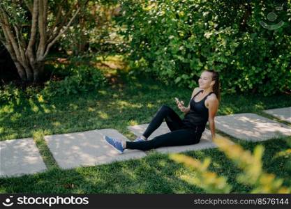 Side view of pleased refreshed woman takes break after active training sits on path in park wears sportsclothes and sneakers listens music via wireless earphones enjoys fresh air and sunny day