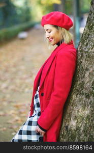 Side view of positive charming female in red coat and beret leaning on tree trunk in autumn park and looking down. Smiling woman in red outfit and skirt leaning on tree