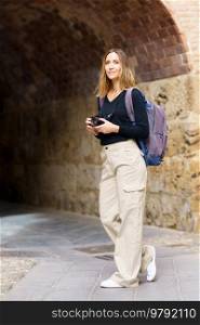 Side view of positive young female tourist with blond hair in casual wear and backpack, using photo camera and smiling while standing near ages stone archway during sightseeing trip. Confident female photographer standing near aged archway during trip