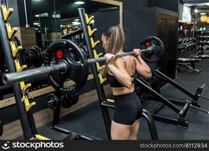 Side view of powerful young female athlete with long ponytail in black sportswear lifting heavy barbell and looking in mirror during workout in gym. Strong young lady exercising with barbell in fitness club