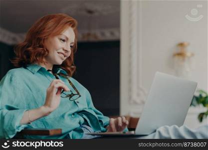 Side view of pretty redhead woman concentrated on remote job, prepares publication for web page, looks gladfully at laptop computer, sits at modern apartment, works remotely, holds eyeglasses
