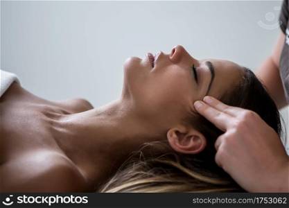 Side view of relaxed woman lying on table in SPA salon and having massage of face keeping eyes closed. Anonymous masseur massaging face skin of woman